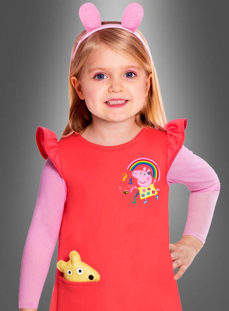Ein kleines Mädchen trägt ein rotes Peppa-Pig-Kleid für Kinder mit einem Peppa-Pig-Motiv, dazu ein rosa Langarmshirt, ein rosa Stirnband mit Hasenohren und ein gelbes Plüschtier in der Tasche – perfekt als Peppa-Wutz-Kostüm für den Kinderkarneval. Im Hintergrund ist ein grauer Farbverlauf zu sehen.