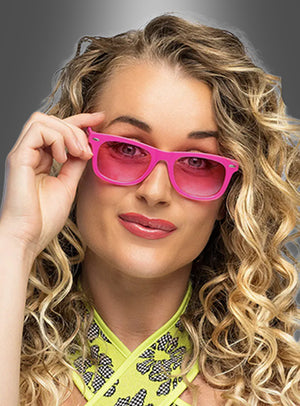 Eine Person mit langen, lockigen blonden Haaren trägt eine rosa „Party Brille“ in verschiedenen Farben sowie ein neon-grün gemustertes Oberteil und blickt mit ausdruckslosem Gesicht vor einem grau verlaufenden Hintergrund in die Kamera.