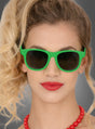 Eine Frau mit welligem blondem Haar trägt eine leuchtend grüne Sonnenbrille im Wayfarer-Stil in verschiedenen Farben, roten Lippenstift und eine rote Perlenkette vor einem grauen Hintergrund.