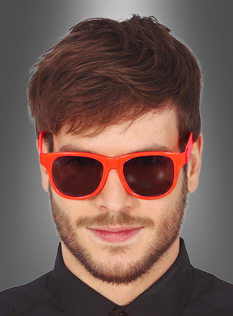 Ein Mann mit braunen Haaren und Bart trägt eine leuchtend rote „Party Brille“ in verschiedenen Farben im klassischen Wayfarer-Stil sowie ein schwarzes Hemd; er blickt mit neutralem Gesichtsausdruck vorwärts vor einem grauen Hintergrund mit Farbverlauf.