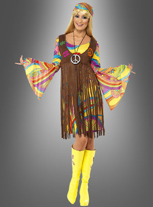 Eine Frau, die das Hippiekostüm „Sunny für Damen“ trägt – bestehend aus einer bunten Fransenweste im Stil der 1960er Jahre, einem passenden Stirnband, einer Halskette mit Peace-Zeichen und gelben Stiefeln –, steht vor einem grauen Hintergrund mit Farbverlauf.