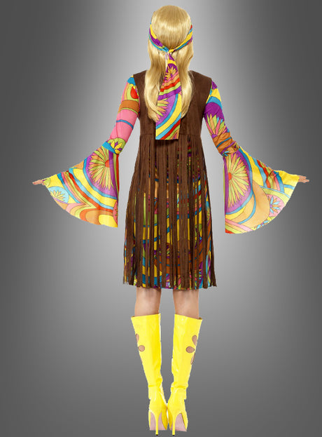 Eine Person steht mit dem Rücken zur Kamera und trägt das „Hippiekostüm Sunny für Damen“ – ein farbenfrohes, psychedelisches Outfit mit ausgestellten Ärmeln, einer braunen Weste mit Fransen, gelb gemusterten Stiefeln und einem passenden Stirnband – vor einem grauen Hintergrund mit Farbverlauf.