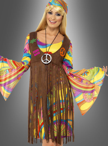 Eine Frau, die das Hippiekostüm „Sunny für Damen“ trägt – mit farbenfrohen Glockenärmeln im Stil der 1960er Jahre, einem Stirnband, einer Weste mit Fransen und einer Halskette mit Peace-Zeichen –, steht vor einem grauen Hintergrund und lächelt.