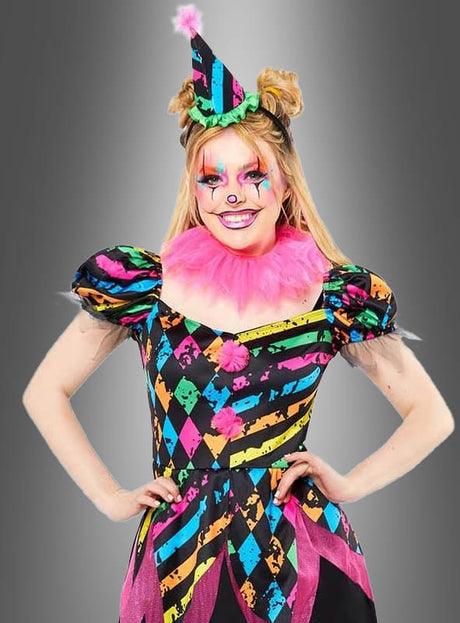 Eine Person, gekleidet in ein farbenfrohes „Zirkus Clown Neon“-Kostüm für Damen, komplett mit passendem Hut, rosa Rüschen und leuchtender Gesichtsbemalung, die vor einem grauen Hintergrund mit Farbverlauf steht.