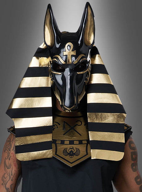 Eine Person, die das „Ägyptischer Gott Anubis Deluxe“-Kostüm trägt – einschließlich einer schwarz-goldenen Maske und eines mit ägyptischen Symbolen verzierten Kopfschmucks –, verkörpert vor einem grauen Hintergrund mit Farbverlauf die geheimnisvolle Aura des Gottes der Toten.