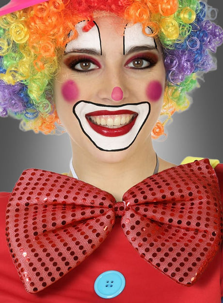 Eine Person lächelt in die Kamera. Sie trägt eine bunte, lockige Clownperücke, weiße Gesichtsbemalung mit übertriebenem Make-up und ein leuchtend rotes Outfit mit einem blauen Knopf, ergänzt durch die „Riesenfliege Karneval“ in verschiedenen Farben als große, gepunktete Fliege.