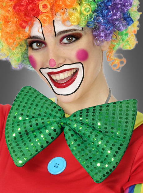 Eine als Clown verkleidete Person mit einer Regenbogenperücke, bunter Gesichtsbemalung und einem roten Kostüm mit blauem Knopf lächelt in die Kamera und trägt dabei die grüne „Riesenfliege“-Karnevalsfliege mit Pailletten.