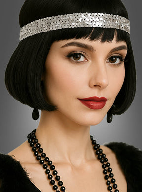Eine Frau mit einem kurzen schwarzen Bob, die ein silbernes Pailletten-Stirnband im Stil der 20er Jahre, roten Lippenstift, eine schwarze Perlenkette und schwarze Ohrringe trägt, posiert vor einem grauen Hintergrund.