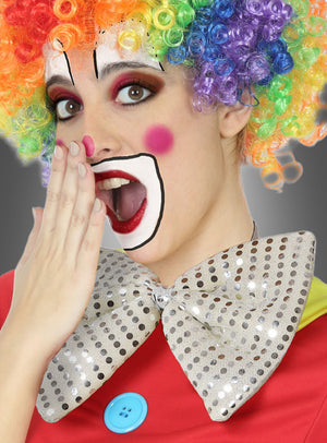Eine Person, die als Clown verkleidet ist – mit einer Regenbogenperücke, bunter Gesichtsbemalung, einem leuchtend roten Kostüm und einer „Riesenfliege“ in verschiedenen Farben –, hält sich in einer überraschten Geste die Hand vor den Mund.