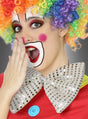 Eine Person, die als Clown verkleidet ist – mit einer Regenbogenperücke, bunter Gesichtsbemalung, einem leuchtend roten Kostüm und einer „Riesenfliege“ in verschiedenen Farben –, hält sich in einer überraschten Geste die Hand vor den Mund.