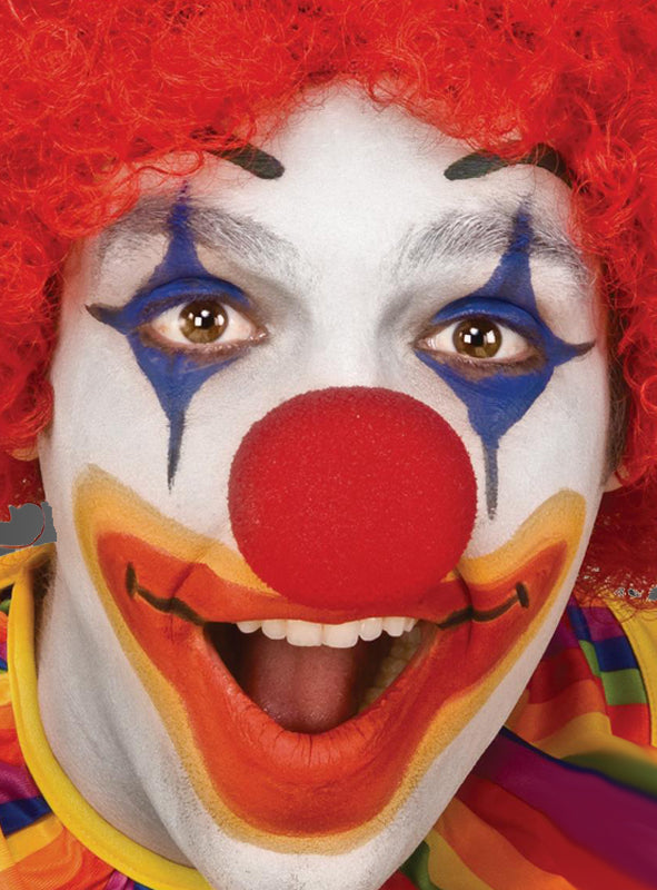 Eine als Clown verkleidete Person mit einer roten Clownnase aus Schaumstoff, einer roten Perücke, weißer Gesichtsfarbe, blauem und orangefarbenem Make-up und einer großen roten Nase, die breit lächelt.