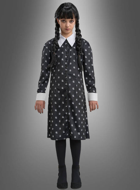 Ein junges Mädchen mit langen schwarzen Zöpfen trägt das „Wednesday Mädchen Kleid schwarz-weiß“ – ein klassisches, von der Addams Family inspiriertes schwarzes Kleid mit weißem Kragen und weißen Manschetten, dazu schwarze Strumpfhosen und Schuhe – und steht vor einem schlichten grauen Hintergrund – die perfekte Wahl für jeden Anlass, bei dem ein Wednesday-Kostüm gefragt ist.