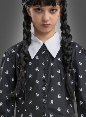 Ein Mädchen mit langen, schwarzen Zöpfen, das das „Wednesday Mädchen Kleid schwarz-weiß“ trägt – ein schwarzes Kleid mit weißem Kragen und Totenkopfmotiven –, steht vor einem grauen Hintergrund und erinnert dabei an den charakteristischen Stil des Wednesday-Kostüms aus der Addams Family.