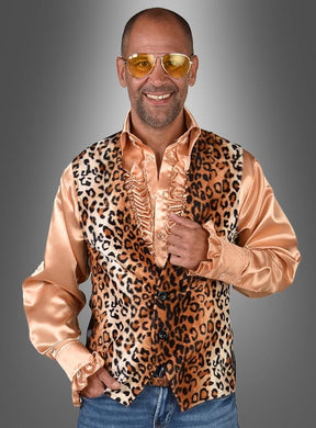 Ein Mann mit einer gelb getönten Brille, einem glänzenden, pfirsichfarbenen Rüschenhemd und einer samtigen Leopardenweste im echten 80er-Jahre-Stil steht vor einem grauen Hintergrund mit Farbverlauf und lächelt.