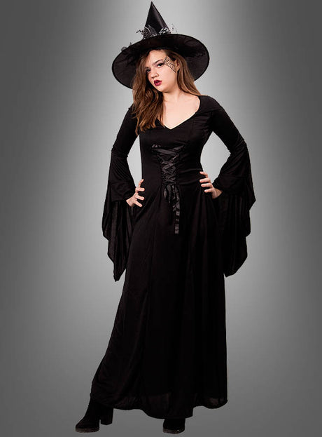Eine Frau, die das Kapuzenkleid „Carmilla“ – ein langes schwarzes Gothic-Kleid – und einen Hexenhut trägt, steht mit einer Hand in die Hüfte gestützt vor einem grauen Hintergrund mit Farbverlauf.