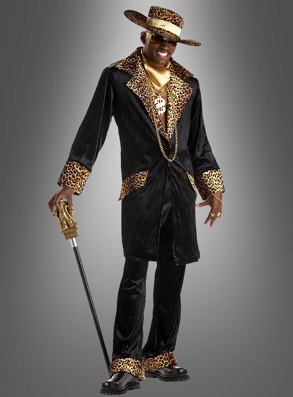 Ein Mann im „Mac Daddy“-Pimp-Kostüm aus den 70ern – bestehend aus einem schwarzen Anzug mit Leopardenmuster-Besatz, einem breitkrempigen Hut, einer Sonnenbrille, goldenen Accessoires und einem Spazierstock mit goldenem Griff – steht vor einem grauen Hintergrund mit Farbverlauf und verkörpert damit perfekt den gewagten Modestil der 70er Jahre.