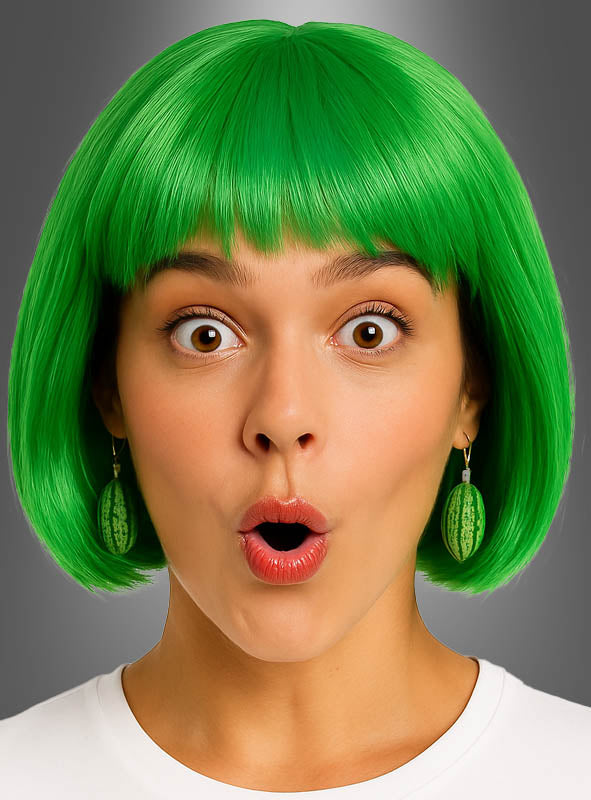 Eine Frau mit einem leuchtend grünen Bob-Haarschnitt, die Ohrringe in Form einer Wassermelonenscheibe oder einer ganzen Frucht sowie weitere verspielte Accessoires mit Fruchtmotiven trägt, öffnet vor einem schlichten grauen Hintergrund mit überraschtem Gesichtsausdruck den Mund.