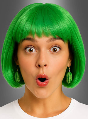 Eine Frau mit einem leuchtend grünen Bob-Haarschnitt, die Ohrringe in Form einer Wassermelonenscheibe oder einer ganzen Frucht sowie weitere verspielte Accessoires mit Fruchtmotiven trägt, öffnet vor einem schlichten grauen Hintergrund mit überraschtem Gesichtsausdruck den Mund.