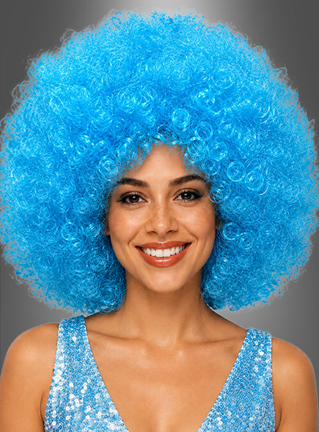 Eine Frau lächelt, während sie eine große, lockige, blaue XXL-Perücke mit der Aufschrift „Riesen Afro Perücken in tollen Farben“ und ein blaues Pailletten-Oberteil trägt; im Hintergrund ist ein neutrales Grau zu sehen.