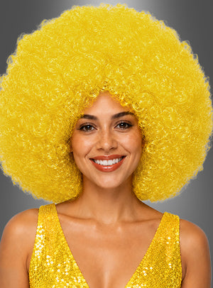 Vor einem grauen Hintergrund lächelt eine Frau in die Kamera. Sie trägt eine XXL-Riesen-Afro-Perücke in leuchtendem Gelb und ein glänzendes gelbes Pailletten-Top.