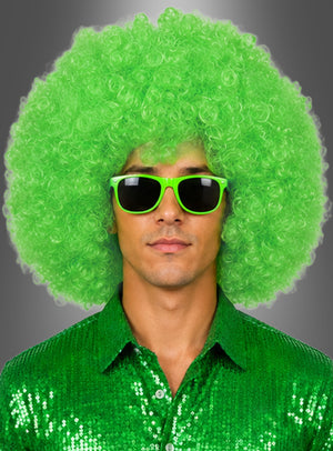 Ein Mann trägt eine XXL-Riesen-Afro-Perücke in leuchtendem, lockigem Grün, dazu eine grüne Sonnenbrille und ein glänzendes grünes Paillettenhemd, vor einem neutralen Hintergrund.