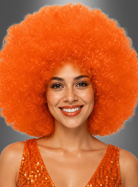 Eine lächelnde Person, die eine orangefarbene XXL-Riesen-Afro-Perücke in tollen Farben und ein orangefarbenes Pailletten-Top trägt und vor einem grauen Hintergrund posiert.