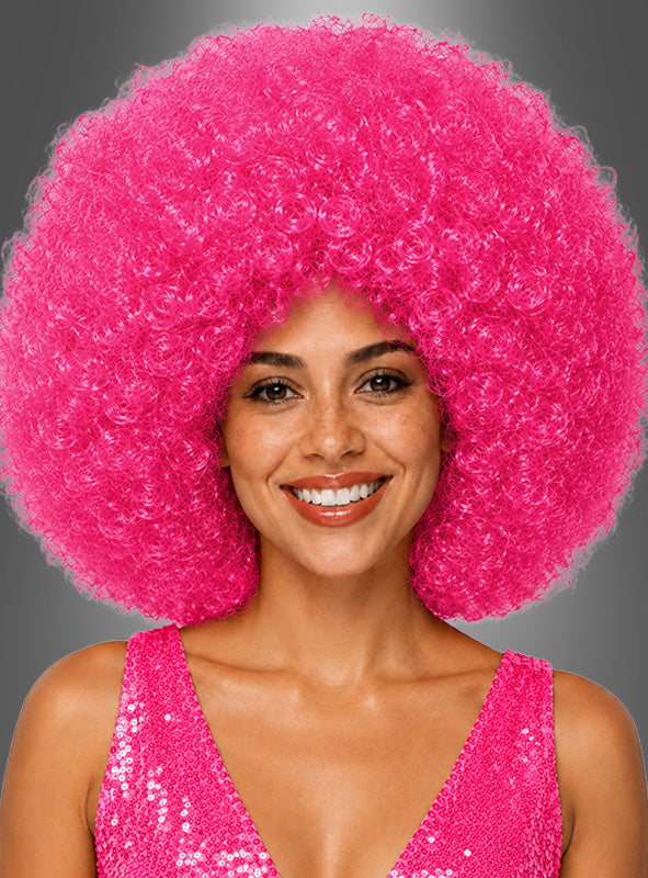 Eine lächelnde Frau trägt eine große, lockige „Riesen-Afro-Perücke in tollen Farben“ in leuchtendem Pink sowie ein glitzerndes, pinkfarbenes, ärmelloses Oberteil vor einem grauen Hintergrund.