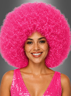 Eine lächelnde Frau trägt eine große, lockige „Riesen-Afro-Perücke in tollen Farben“ in leuchtendem Pink sowie ein glitzerndes, pinkfarbenes, ärmelloses Oberteil vor einem grauen Hintergrund.