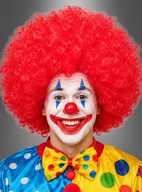 Eine Person in einem bunten Clownskostüm, die eine leuchtend rote Riesen-Afro-Perücke in Größe XXL aus der Kollektion „Riesen-Afro-Perücken in tollen Farben“ trägt, mit weißer Gesichtsbemalung, einer roten Nase und einer großen, bunten Fliege mit Polka-Punkten, die in die Kamera lächelt.