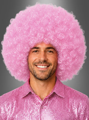 Ein Mann lächelt, während er eine riesige Afro-Perücke in Rosa (XXL, groß und lockig) sowie ein glänzendes rosa Paillettenhemd trägt, vor einem grauen Hintergrund mit Farbverlauf.