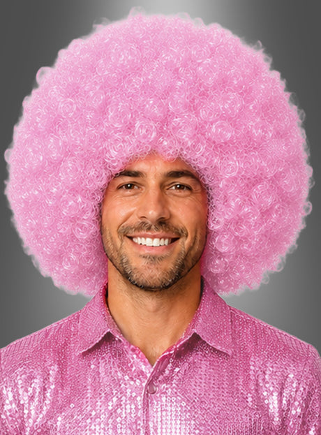 Ein Mann lächelt, während er eine riesige Afro-Perücke in Rosa (XXL, groß und lockig) sowie ein glänzendes rosa Paillettenhemd trägt, vor einem grauen Hintergrund mit Farbverlauf.