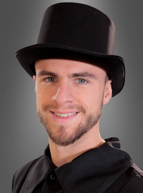 Ein junger Mann mit kurzem Bart und Schnurrbart lächelt vor einem schlichten grauen Hintergrund in die Kamera; er trägt den „Deluxe Satin Zylinder schwarz“ und ein schwarzes Outfit.