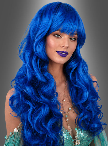 Eine Frau mit langen, welligen, leuchtend blauen Haaren, die als „Lockenperücke mit Pony Melli“ frisiert sind, und blauem Lippenstift, die ein grünes Outfit mit Perlenverzierungen im echten Karnevalskostüm-Flair trägt, posiert vor einem grauen Hintergrund mit Farbverlauf.