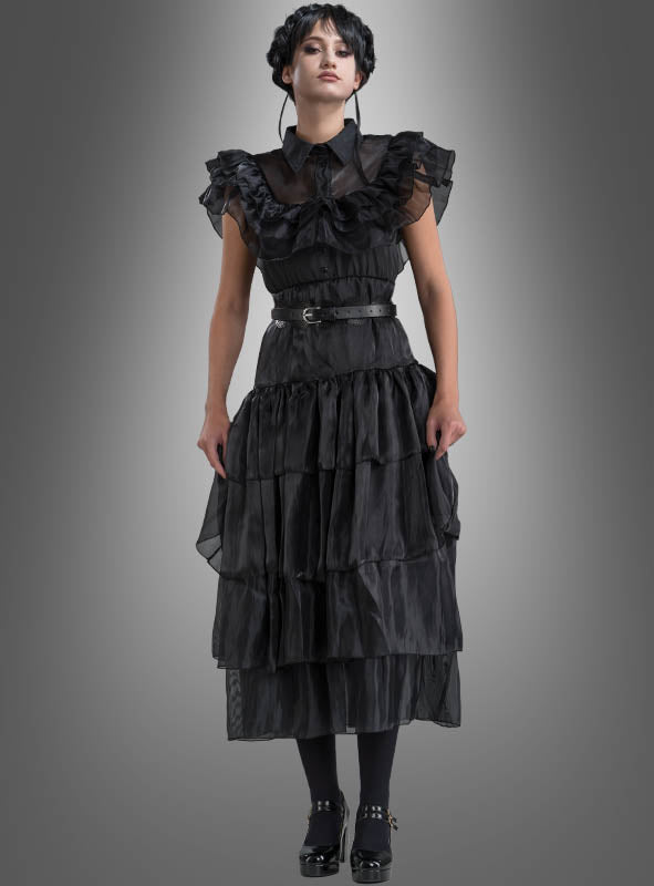 Eine Frau blickt nach vorne und trägt das „Wednesday Damen Ballkleid“ – ein mehrlagiges schwarzes Rüschenkleid mit transparenten Ärmeln –, kombiniert mit einem schwarzen Gürtel, schwarzen Strumpfhosen und schwarzen Schuhen vor einem grauen Hintergrund mit Farbverlauf, der an den ikonischen Look der Addams Family erinnert.