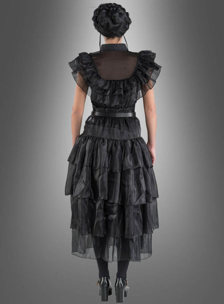 Eine Person steht mit dem Rücken zur Kamera und trägt das „Wednesday Damen Ballkleid“ – ein mehrlagiges schwarzes Rüschenkleid –, kombiniert mit schwarzen Leggings und schwarzen Stöckelschuhen, vor einem grauen Hintergrund mit Farbverlauf.