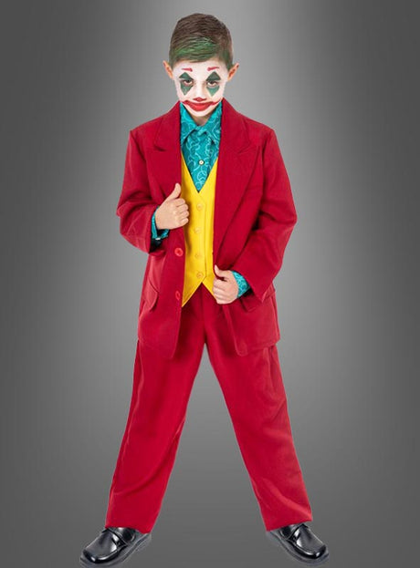 Ein Kind, das den „Roten Joker-Anzug für Kinder“ trägt – bestehend aus einem roten Anzug, einer gelben Weste, einem blaugrünen Hemd und einer Clown-ähnlichen Gesichtsbemalung –, steht vor einem grauen Hintergrund.