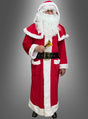 Eine Person, die einen Weihnachtsmann-Mantel mit Stiefelüberzügen trägt, einen weißen Bart und ein rotes Plüschkostüm hat und eine kleine goldene Glocke in der Hand hält, während sie vor einem grauen Hintergrund steht.