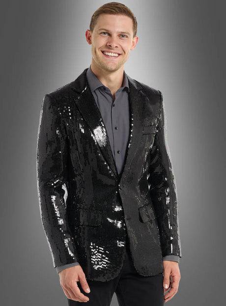 Ein Mann steht vor einem grauen Hintergrund mit Farbverlauf, trägt eine schwarze Herrenjacke mit Pailletten über einem Hemd mit Knöpfen und lächelt in die Kamera.