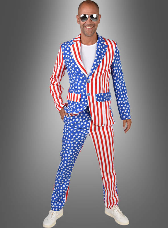 Ein Mann steht vor einem grauen Hintergrund und lächelt. Er trägt das „USA Anzug Amerika“-Kostüm mit auffälligem „Stars & Stripes“-Muster der amerikanischen Flagge, dazu ein weißes T-Shirt, weiße Turnschuhe und eine Sonnenbrille.