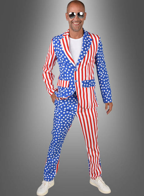 Ein Mann steht vor einem grauen Hintergrund und lächelt. Er trägt das „USA Anzug Amerika“-Kostüm mit auffälligem „Stars & Stripes“-Muster der amerikanischen Flagge, dazu ein weißes T-Shirt, weiße Turnschuhe und eine Sonnenbrille.