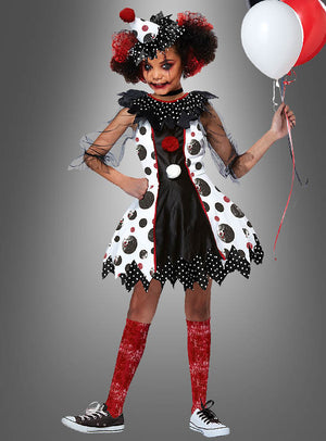 Ein Kind, das das Clown-Kleid-Mädchenkostüm „Pippa“ trägt – ein schwarz-weiß-rot gepunktetes Narrenkostüm –, hält rote, weiße und schwarze Luftballons in der Hand und steht vor einem grauen Hintergrund.