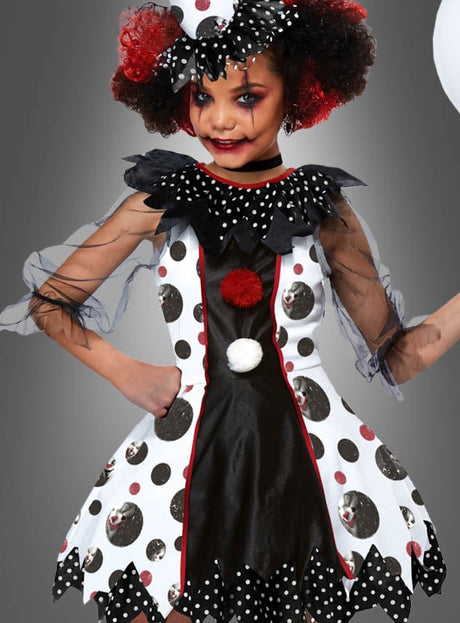 Ein Kind, das das Clown-Kleid „Mädchen-Kostüm Pippa“ trägt – mit schwarzen, weißen und roten Polka-Punkten, auffälligem Make-up und einem Rüschenkragen –, posiert mit einer Hand in die Hüfte gestützt vor einem grauen Hintergrund. Diese „böse Clown“-Verkleidung verleiht dem Look eine besonders unheimliche Note.