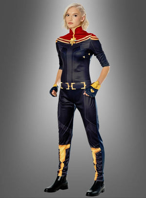 Eine Person steht vor einem grauen Hintergrund und trägt das Captain-Marvel-Kostüm für Damen, das sich durch auffällige rote, blaue und goldene Farben auszeichnet, dazu schwarze Stiefel und kurzes blondes Haar – ein kraftvolles Marvel-Superheldinnen-Kostüm für Damen, das vom Kostüm-Ensemble der Avengers inspiriert ist.