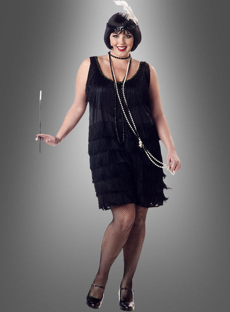 Eine Frau, die das schwarze Charleston-Kleid in Größe XXL mit Fransen trägt, ergänzt durch Netzstrümpfe, eine Perlenkette und ein mit Federn verziertes Stirnband, und eine Zigarettenspitze in der Hand hält, steht vor einem grauen Hintergrund und verkörpert die Eleganz der Mode der 20er Jahre.