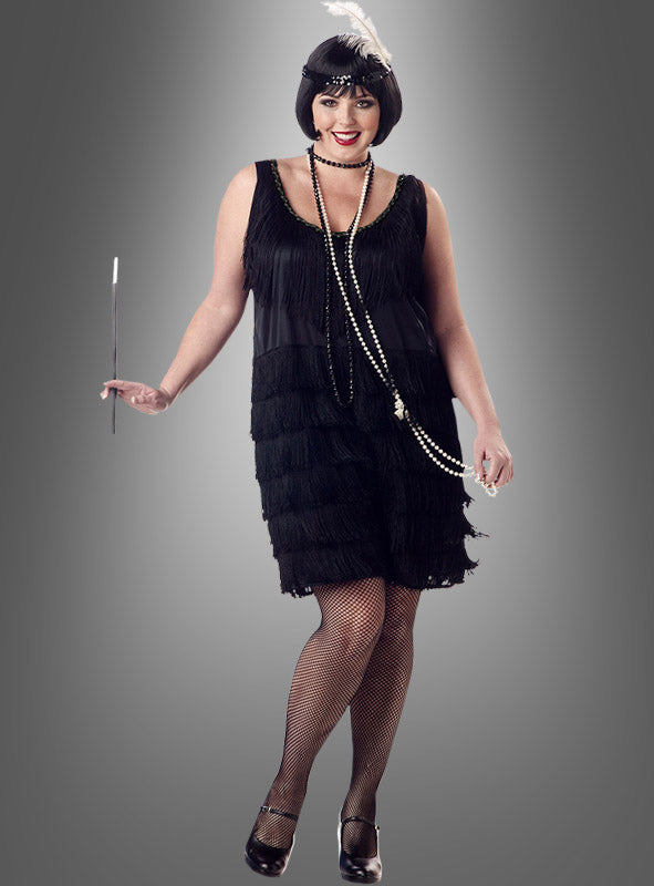 Eine Frau, die das schwarze Charleston-Kleid in Größe XXL mit Fransen trägt, ergänzt durch Netzstrümpfe, eine Perlenkette und ein mit Federn verziertes Stirnband, und eine Zigarettenspitze in der Hand hält, steht vor einem grauen Hintergrund und verkörpert die Eleganz der Mode der 20er Jahre.