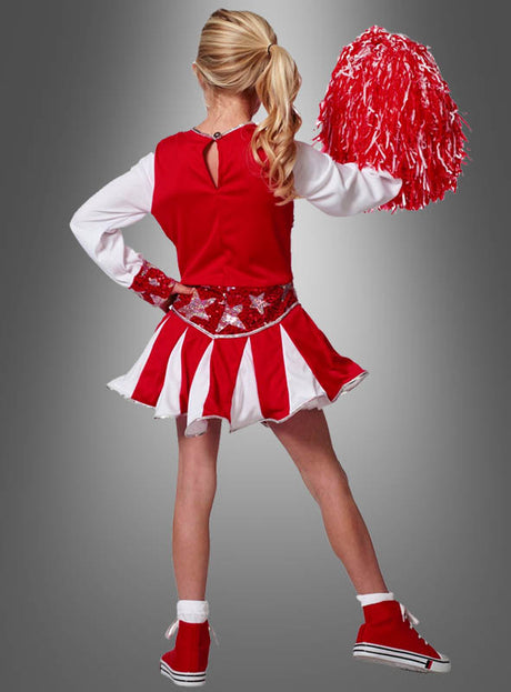 Ein Mädchen, das ein „Cheerleader-Kostüm für Mädchen“ in Rot und Weiß trägt, steht mit dem Rücken zur Kamera und hält vor einem grauen Hintergrund einen roten Pompon in der rechten Hand.