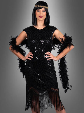 Eine Frau, die das „Edles Gatsby Kleid Daisy“ trägt – ein schwarzes, mit Perlen besticktes Flapper-Kleid, das von der Mode der 20er Jahre inspiriert ist und mit einer schwarzen Federboa sowie einem Stirnband ergänzt wird –, steht mit den Händen in den Hüften vor einem grauen Hintergrund mit Farbverlauf.