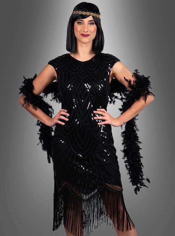Eine Frau, die das „Edles Gatsby Kleid Daisy“ trägt – ein schwarzes, mit Perlen besticktes Flapper-Kleid, das von der Mode der 20er Jahre inspiriert ist und mit einer schwarzen Federboa sowie einem Stirnband ergänzt wird –, steht mit den Händen in den Hüften vor einem grauen Hintergrund mit Farbverlauf.