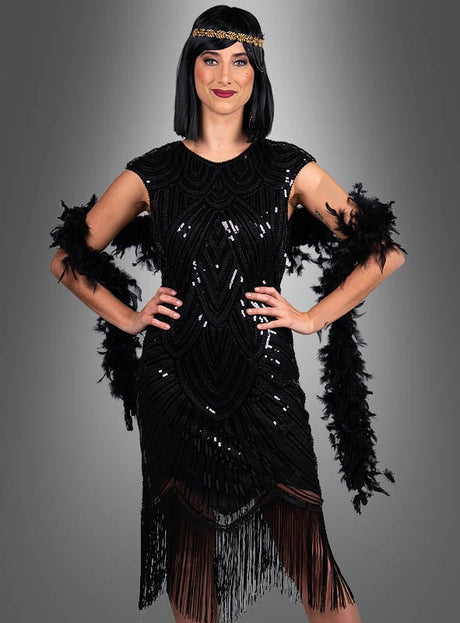 Eine Frau trägt das „Edles Gatsby Kleid Daisy“, ein schwarzes Flapper-Kleid mit Fransen, das an die Mode der 20er Jahre erinnert, kombiniert mit einer schwarzen Federboa und einem mit Perlen verzierten Stirnband; sie steht vor einem grauen Hintergrund mit Farbverlauf – und verkörpert damit perfekt den ikonischen Stil von „Der große Gatsby“.