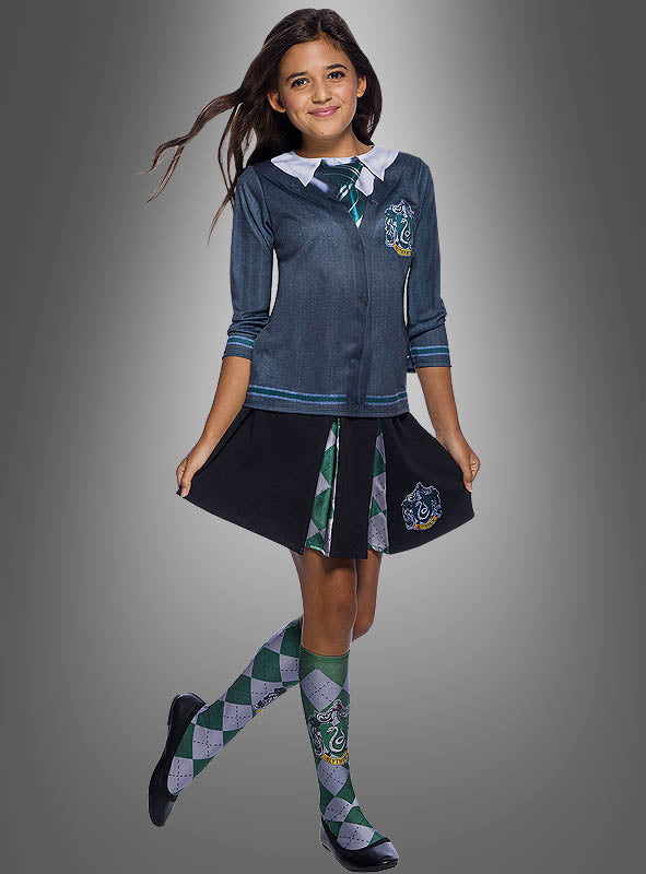 Ein Mädchen, das das Slytherin-Shirt aus „Harry Potter“ für Kinder trägt – dazu eine grüne Krawatte, einen Faltenrock und karierte Socken –, steht vor einem grauen Hintergrund.